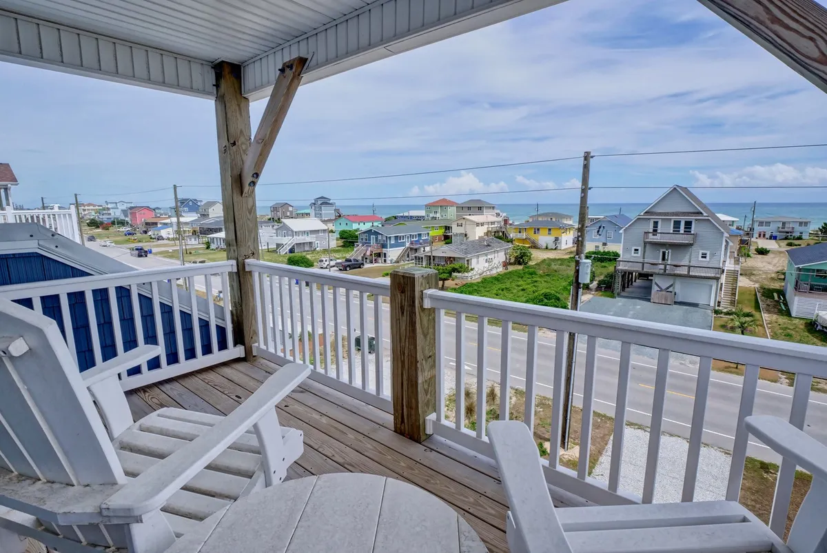 A spacious wooden deck overlooks the ocean, framed by neighboring houses and lush greenery. The deck is set up for quiet relaxation, perfect for enjoying the coastal breeze or watching the sunset over the water.