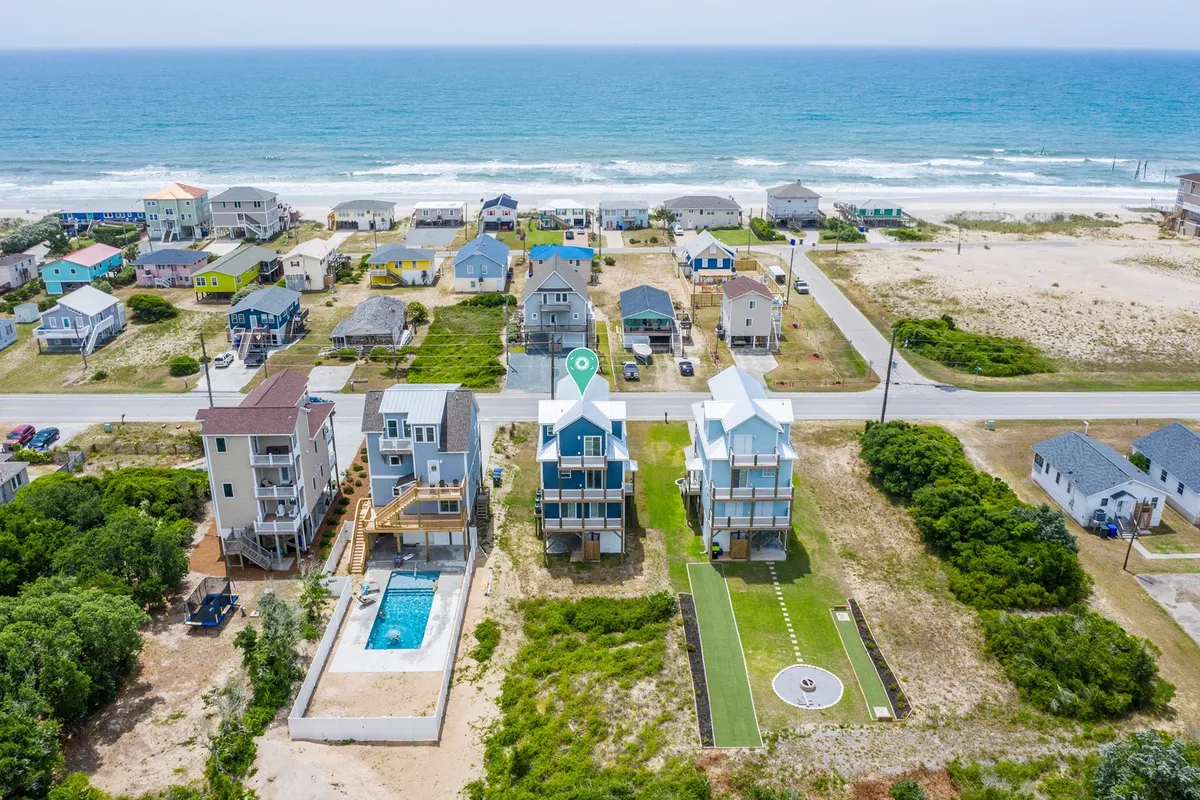 This image captures a picturesque beachfront setting with colorful houses, a sparkling swimming pool, and direct access to the sandy shore. Families can be seen enjoying the poolside as well as the beach beyond, under a relaxed sunset sky.