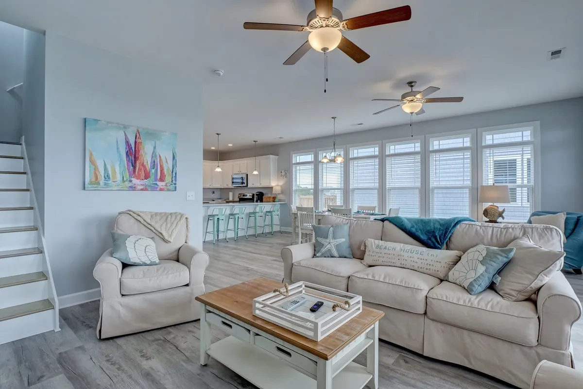 A comfortable living area with a plush sofa, decorative pillows, a wooden coffee table, colorful wall art, and a kitchen with bar stools, all bathed in natural light streaming through large windows. The open layout indicates easy flow from indoor to outdoor spaces.