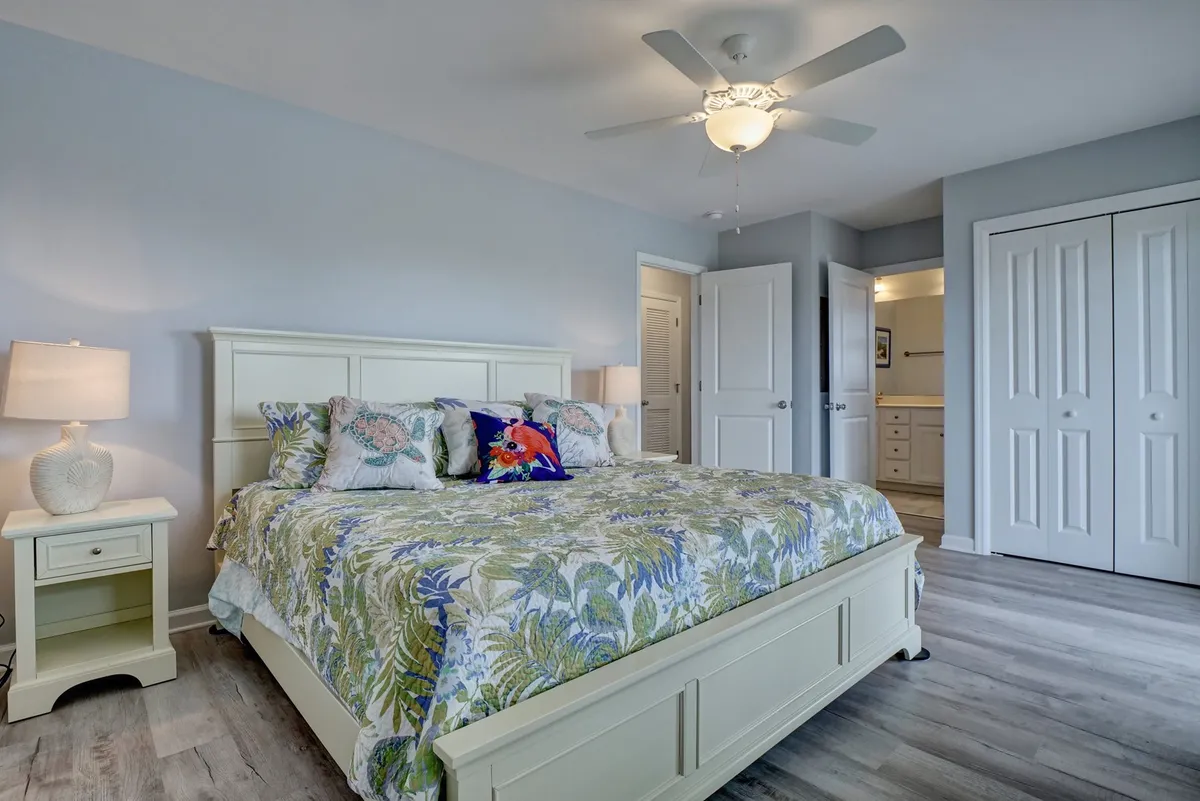 A tranquil bedroom featuring a bed with colorful bedding, a nightstand with a lamp, and a ceiling fan overhead. Natural light softly filters through the windows, creating a cozy and restful ambiance, ideal for relaxation or reading.