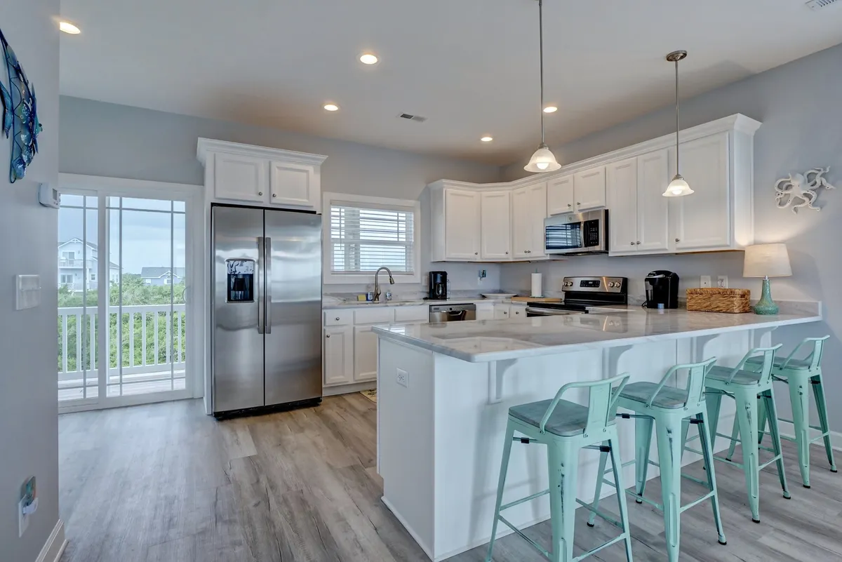 A modern kitchen features a spacious island with bar stools, stainless steel appliances, and expansive windows letting in sunlight. The layout encourages both cooking and socializing, with ample space for gathering around the island or preparing meals as a group.