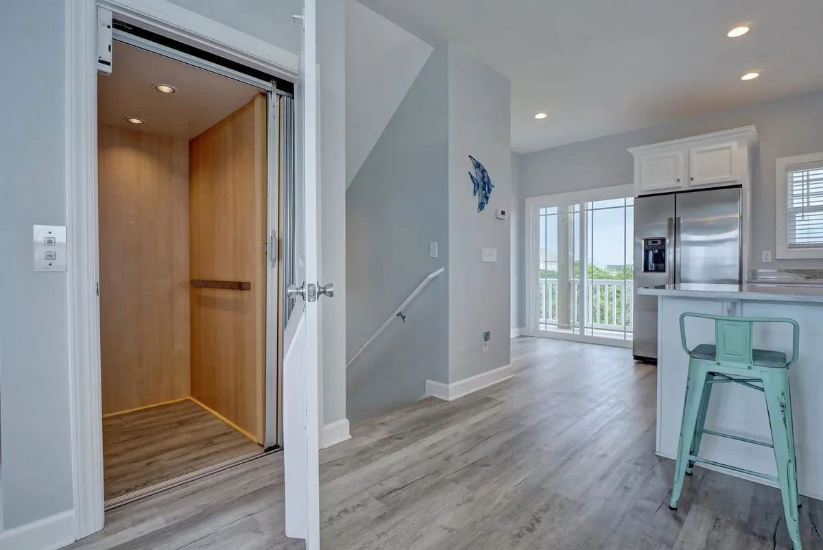 The image shows a modern and inviting interior featuring an elevator adjacent to a stylish kitchen area, with large windows allowing natural light to fill the space. The kitchen is equipped with contemporary appliances and comfortable seating, clearly illustrating accessibility and comfort.