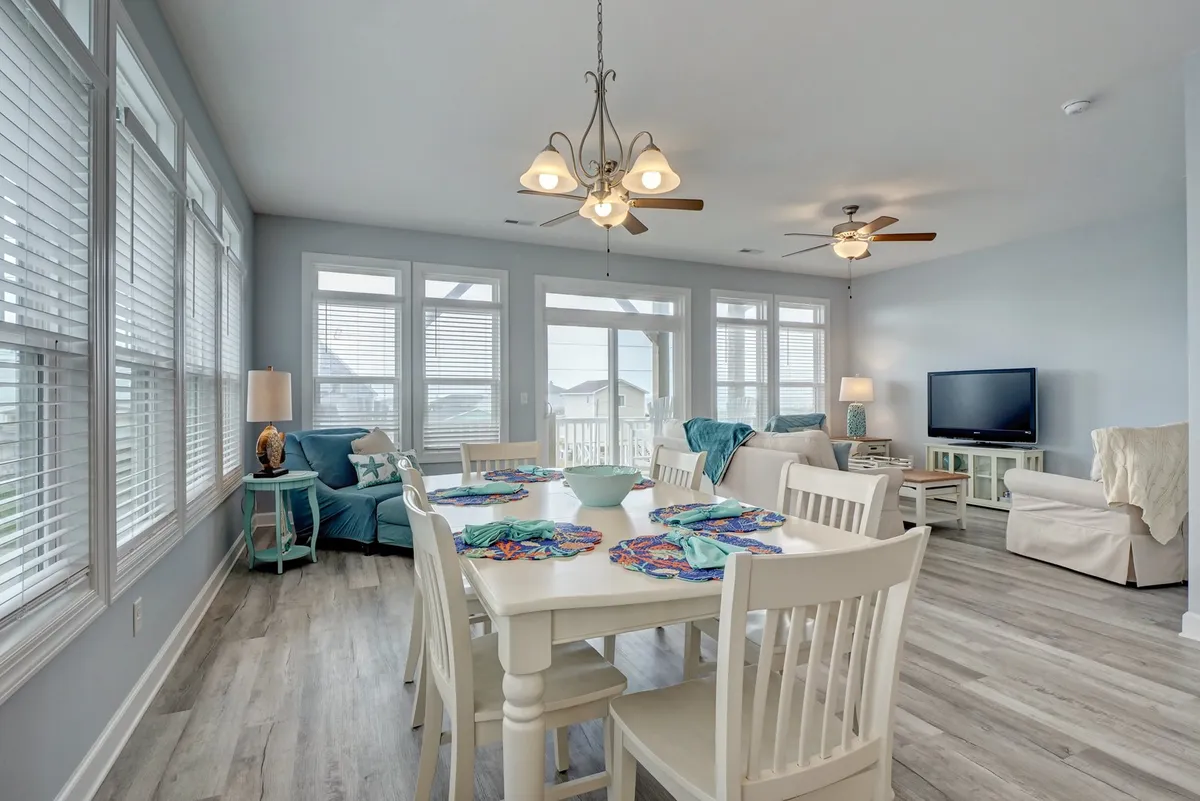 This scene includes a dining table with colorful placemats, surrounded by large windows that allow abundant natural light. Beyond the table, a comfortable seating area with blue cushions and a stylish chandelier create an inviting atmosphere for meals, games, or conversation.
