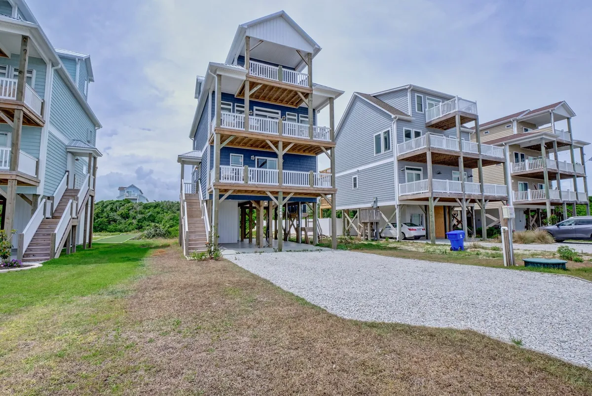This image features the three-story house at 2669 Island Drive, perched on stilts with a visible gravel driveway and neighboring homes, setting a welcoming scene for family arrivals. The structure and elevation suggest accessibility features and space for all ages.