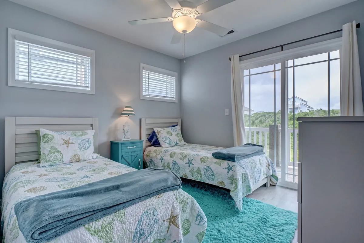 A calm and inviting twin bedroom with two beds, a turquoise nightstand between them, and a large window draped with curtains. The atmosphere is serene and sunlit, offering a peaceful retreat for family or friends.