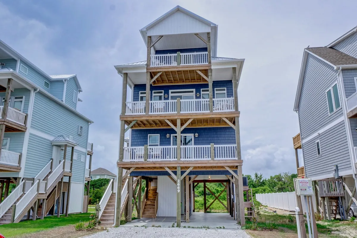 The image showcases a welcoming three-story beach house with multiple spacious balconies, supported by rustic wooden beams. The home's generous outdoor spaces invite guests to gather, relax, and soak in the coastal ambiance. The elevated structure hints at sweeping views—perfect for sunset watching and enjoying cool ocean breezes.