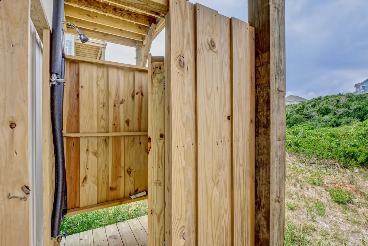 This exterior image features a welcoming wooden outdoor shower nestled beside the home, surrounded by lush greenery. The scene invites guests to rinse off after beach activities, with a casual and inviting mood that seamlessly ties to outdoor relaxation and play.